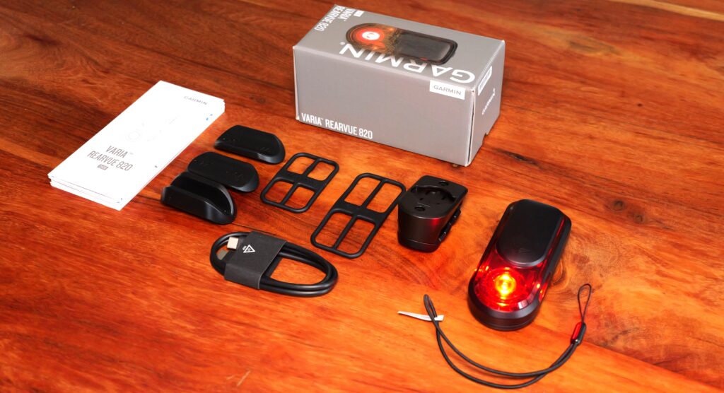 Garmin RearVue 820 Package Contents on a Wooden Table: Rear Light, Mount, Rubber Straps, Spacers, USB Cable, Safety Tether, Manual and Packaging.